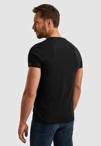 Mann mit kurzen braunen Haaren, der ein schwarzes kurzärmliges T-Shirt und blaue Jeans trägt, mit dem Rücken zu einem einfarbigen hellgrauen Hintergrund.