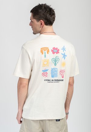Mężczyzna w kremowej koszulce z kolorowymi, tropikalnymi motywami roślinnymi i napisem „Living in Paradise Kaotiko Society” na plecach.