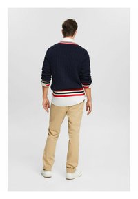Maglione navy a trecce con dettagli a strisce rosse e bianche, indossato sopra una camicia bianca con colletto, abbinato a pantaloni khaki e sneakers bianche.
