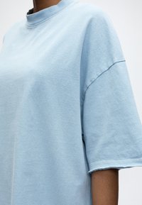 Άτομο που φοράει ανοιχτό μπλε κοντομάνικη μπλούζα με στρογγυλή λαιμόκοψη, εμφανίζοντας το πάνω μέρος του κορμού και το χέρι σε απλό φόντο.