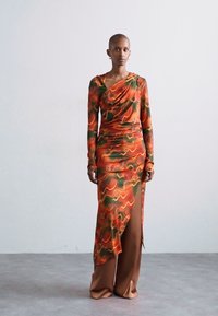 Long-sleeve dress in colorful swirling pattern of orange and green, draped neckline, side slit, paired with smooth brown wide-leg pants.