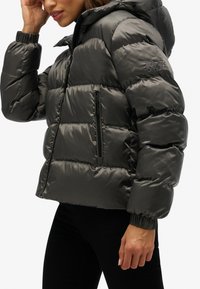 Veste matelassée rembourrée en gris anthracite brillant avec capuche amovible, poignets élastiques, poche poitrine zippée et design matelassé horizontal.