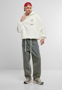 Sweatshirt blanc oversize avec poche avant, arborant des logos textuels noirs. Associé à un jean ample délavé et des baskets de sport grises. Casquette rouge.