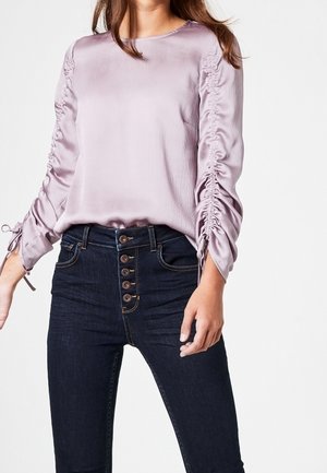 Mujer con blusa de manga larga arrugada de color púrpura claro metida en unos vaqueros de talle alto de color azul oscuro con cierre de botones, sobre un fondo blanco.