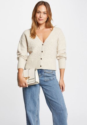 Beige gestrickte Strickjacke mit V-Ausschnitt, gerippten Bündchen und fünf Knöpfen. Kombiniert mit hoch geschnittenen blauen Jeans und einer cremefarbenen Handtasche mit goldenen Akzenten.