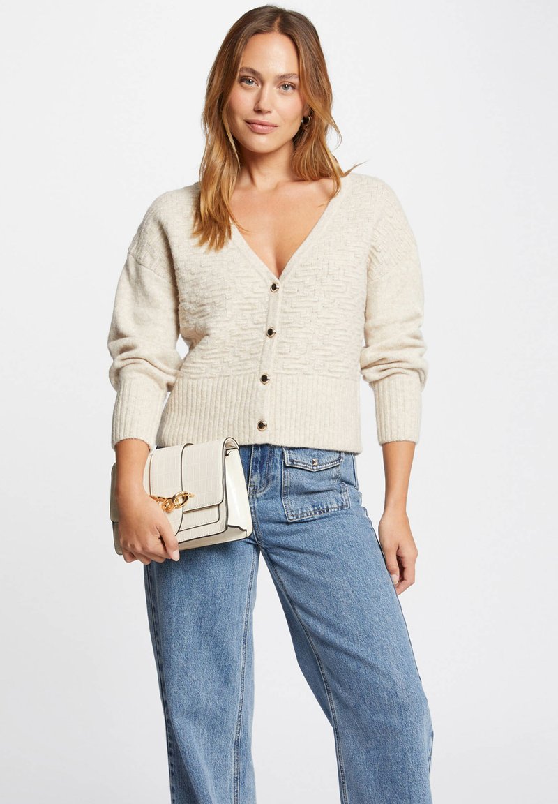 Beige stickad cardigan med V-ringning, ribbade ärmar och fem knappar. Matchad med högmidjade blå jeans och en krämfärgad handväska med guldfärgad accent.