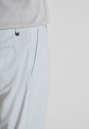 Lichtgrijze, gestructureerde pantalon gedragen met een ingestopte beige blouse, met de nadruk op de taille- en zakgebieden tegen een effen achtergrond.