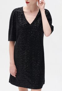 Vestido negro de lentejuelas con escote en V, mangas cortas y un corte holgado. La tela presenta una superficie texturizada creada por las lentejuelas.