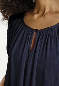 Blouse bleu marine présentant une texture plissée douce, un décolleté rond avec un détail œillet, et une fermeture par bouton au col pour un accent supplémentaire.