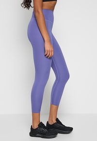 Fioletowe spodnie sportowe z wysokim stanem, wykonane z elastycznego materiału. Charakteryzują się gładką teksturą i sięgają do połowy łydki. W połączeniu z czarnymi trampkami.