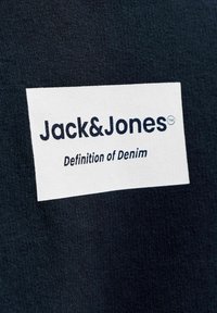 Valge ristkülikukujuline plaastrik tume kanga peal, millel on "Jack&Jones" logo ja musta tekstiga loosung "Definition of Denim".