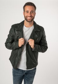 Giubbotto da motociclista in pelle verde scuro con dettagli in zip argentati, alette e una texture liscia, indossato sopra una t-shirt bianca e jeans blu.