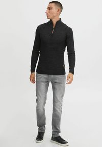 Svart zip-neck genser med ribbet tekstur, tettsittende silhuett og lange ermer, kombinert med lysegrå jeans og svarte sneakers.