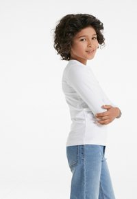 Weißes langärmliges Shirt mit Rundhalsausschnitt, kombiniert mit hellblauen geraden Jeans. Kind steht seitlich mit verschränkten Armen.