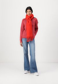 Rød ribbestrikket genser kombinert med et langt rødt skjerf; utsvingte lyseblå jeans og hvite joggesko fullfører antrekket. Minimal bakgrunn.