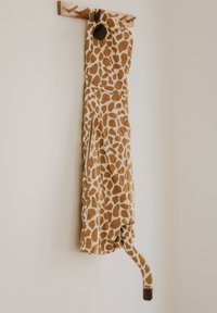 Apģērbs ar žirafes rakstu, kurā dominē brūnas un krēmkrāsas toņi, ar mīkstu audumu un asti apakšā. Karājas uz koka āķa.