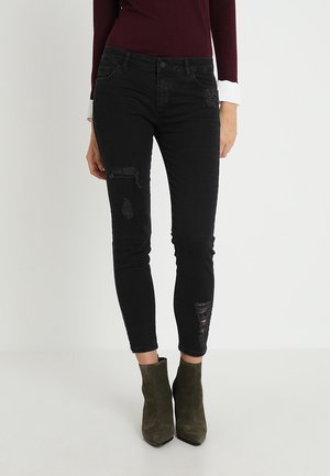 Jeans skinny de mezclilla negra con detalles desgastados. Los jeans se combinan con botines de ante verde oliva, lo que resalta una silueta estilizada.
