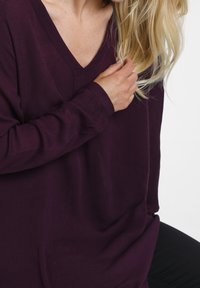 Långärmad V-ringad topp i djup lila, gjord av lättviktig tyg med en mjuk textur. Har en avslappnad passform och diskreta sidoflikar.