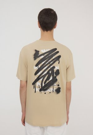 T-shirt in cotone beige con un design spray nero e bianco sulla schiena, caratterizzato da gocce e forme astratte. Maniche corte e vestibilità standard.