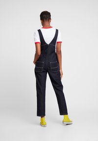 Mono de mezclilla oscuro con tirantes, que presenta dos bolsillos traseros y costuras blancas. Combinado con una camiseta blanca con acentos rojos y zapatos amarillos.