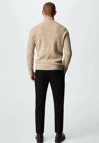 Uomo che indossa un maglione a collo alto rigato beige, pantaloni neri e stivali marroni scuri, in piedi di spalle contro uno sfondo chiaro e uniforme.