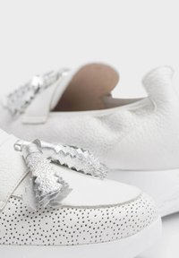 Las zapatillas de cuero blancas presentan una superficie texturizada y un lazo metálico plateado con bordes en zigzag, montadas sobre una suela blanca.