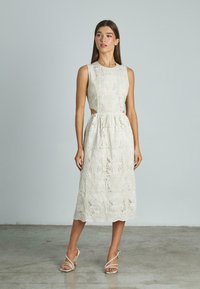 Abito midi in pizzo color crema con lati scoperti e vita aderente, caratterizzato da intricate fantasie floreali e una gonna svasata. Tacchi nudi con cinturini.