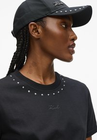 Sort T-shirt med rund hals, prydet med sølvnitter og et broderet logo. Kombineret med en matchende sort kasket med en kant med nitter.