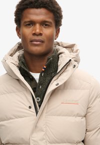 Beige polstret jakke med hætte, der har lynlås og knaplukning. Det indre lag er en mørkegrøn strikket sweater med knapper.