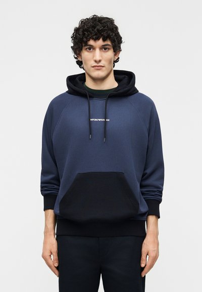 Fiatal férfi göndör sötét hajjal, tengerészkék és fekete kapucnis pulóvert visel, amely elülső zsebbel és húzózsinórokkal rendelkezik.