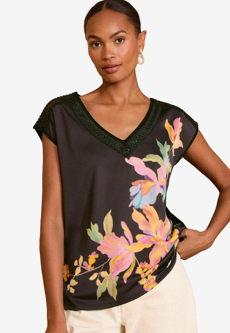 Melnas īsām piedurknēm blūze ar V-veida izgriezumu, kurai ir krāsaini ziedu raksti rozā, dzeltenā un zilā krāsā, un strukturēts melns apmales gar izgriezumu.