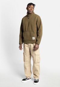 Pull en polaire vert olive avec demi-fermeture éclair et poche avant, associé à un pantalon cargo beige et des baskets noires. Tissu texturé, coupe décontractée.
