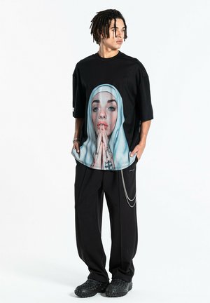 Joven con rastas que lleva un conjunto negro de gran tamaño y una camiseta gráfica con una mujer rezando con tatuajes y piercings.