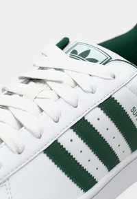 Witte leren sneaker met groene rubberen strepen, witte veters, en een groen logo op de tong. Gestructureerde accenten versieren de zijkant van de schoen.