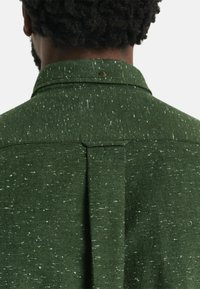 Groene gestructureerde blouse met een gespikkeld patroon, omgeslagen kraag en een plooi aan de achterkant met een zichtbare knoopdetail.