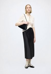 Cream short-sleeve sweater with gold buttons, paired with a black satin skirt. Model holds a black bag, wearing beige socks and black flats.