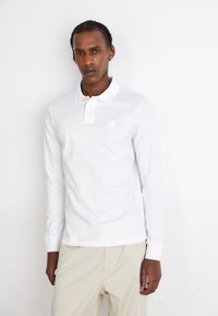 Cămașă polo albă cu mânecă lungă, guler cu nasturi și un mic logo pătrat. Textură netedă; asortată cu pantaloni bej deschis.