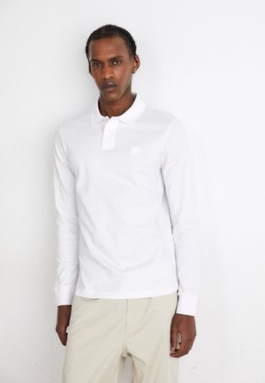 Weißes Langarm-Polohemd mit Knopfkragen und kleinem quadratischen Logo. Glatte Textur; kombiniert mit hellbeigen Hosen.