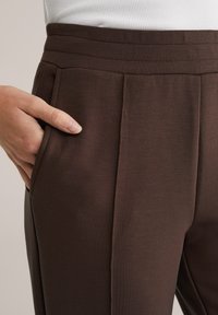 Kéz egy barna, magasderekú nadrág zsebében, varrási részlettel, amelyet fehér bordázott felsővel viselnek.