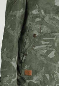 Grön jacka med camouflagemönster i texturerad tyg, med en dragkedjeficka, knappstängning och en läderetikett med varumärkets logotyp.