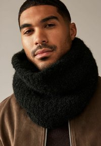Chunky black knitted scarf with a soft, textured finish, worn loosely around the neck. Background is neutral, highlighting the scarf.