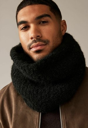 Chunky black knitted scarf with a soft, textured finish, worn loosely around the neck. Background is neutral, highlighting the scarf.