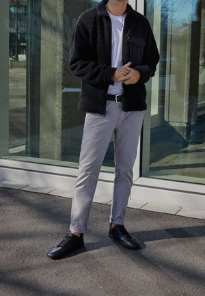 Mies seisoo lasirakennuksen ulkopuolella musta fleece-takki, valkoinen paita, harmaat housut ja mustat tennarit päällään, kädet ristissä.