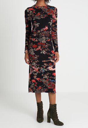 Vrouw draagt een zwarte midi-jurk met rood en blauw bloem- en vogelpatroon, gecombineerd met donkergroene veterlaarsjes met blokhakken.