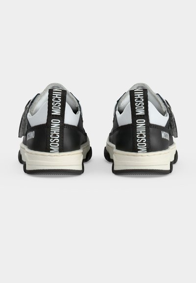 Ζευγάρι ασπρόμαυρα sneakers Moschino, ορατά από πίσω με μπρελόκ με το λογότυπο και παχιά λευκή σόλα σε μια απλή φόντο.