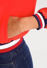 Röd pullover med ballongärmar, med blåvita randiga mudd och midjeband. Bärs över mörkblå denim med en slät yta.