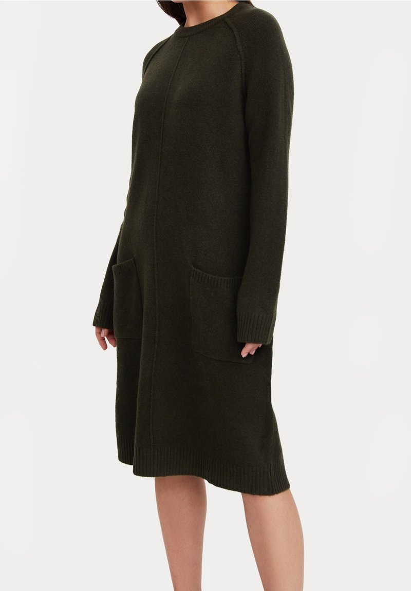 Femme portant une robe pull en maille vert foncé, manches longues, longueur genou, avec deux poches avant, sur un fond clair uni.