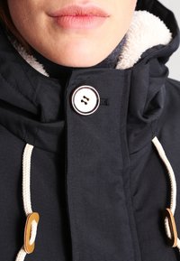 Marinblå jacka med en huva fodrad med fuskpäls, som har en stor vit knapp och krämfärgade snoddar. Strukturerad tyg med detaljerad sömnad.