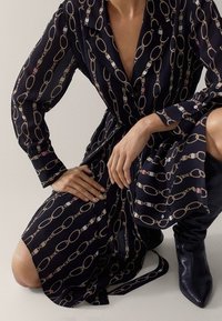 Laivansinis pitkähihaisellinen mekko, jossa on ketjumainen kuvio, varustettuna kauluksella, vyöllä ja lonnelaajuisella siluetilla. Yhdistettynä mustiin polvimittaisiin saappaisiin.