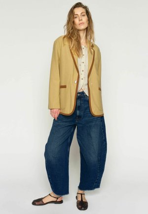 Žena v béžovom saku s hnedým lemovaním, svetlá blúzka s vzorom, široké modré džínsy a čierne pletené sandále, stojaca pred jednofarebným pozadím.